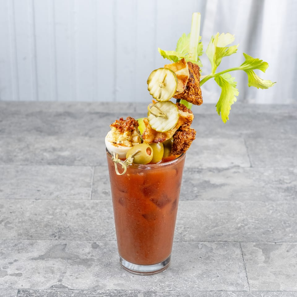 Bloody Mary cocktail topped with fried chicken, pickles, olives, deviled egg, celery, and garnishes on a stone table.