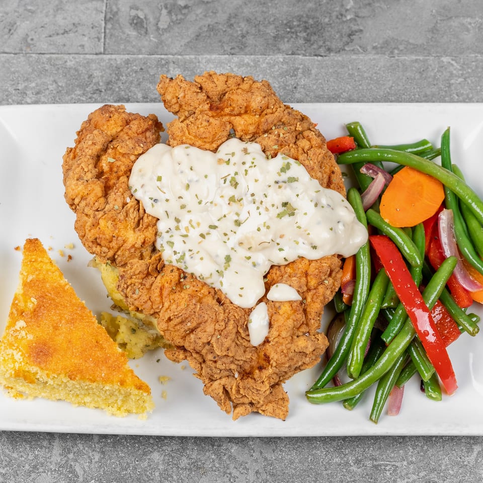 Plate of crispy fried chicken topped with creamy gravy, served with cornbread and colorful green bean vegetables.