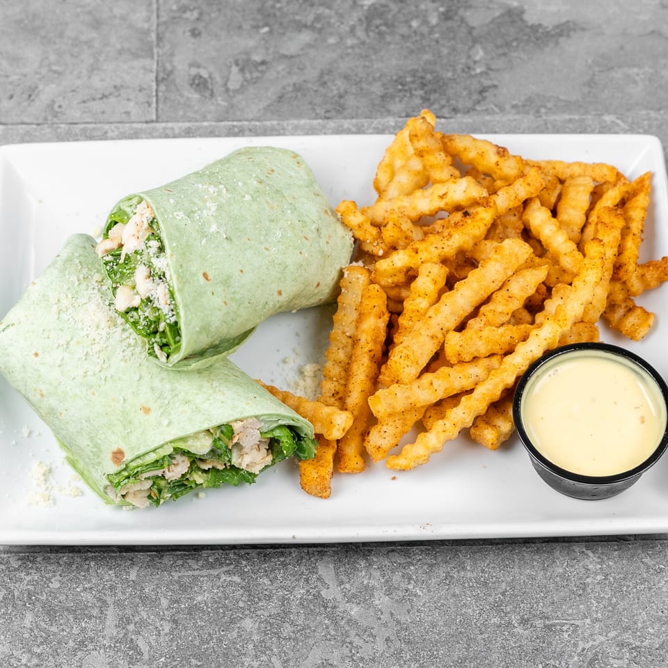 Chicken Caesar wrap with leafy greens and crinkle-cut seasoned fries served with creamy dipping sauce on a white plate.
