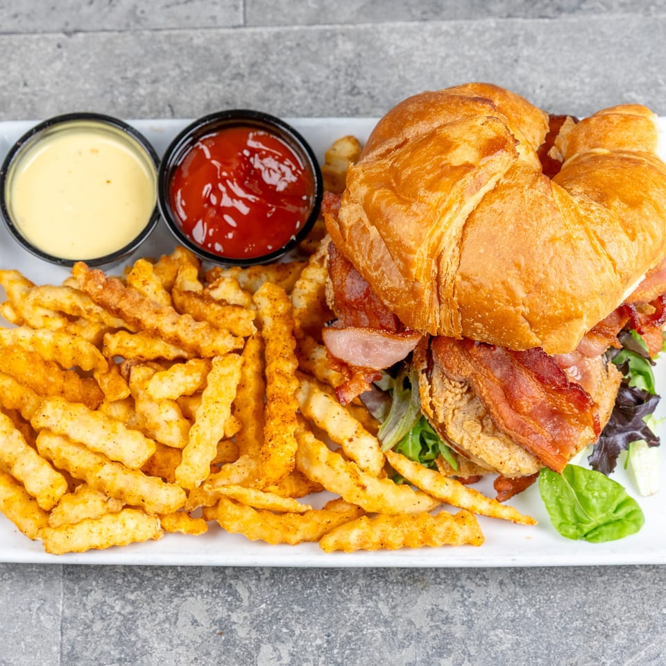 Crispy chicken and bacon croissant sandwich with seasoned crinkle-cut fries, cheese sauce, and ketchup on a white plate.