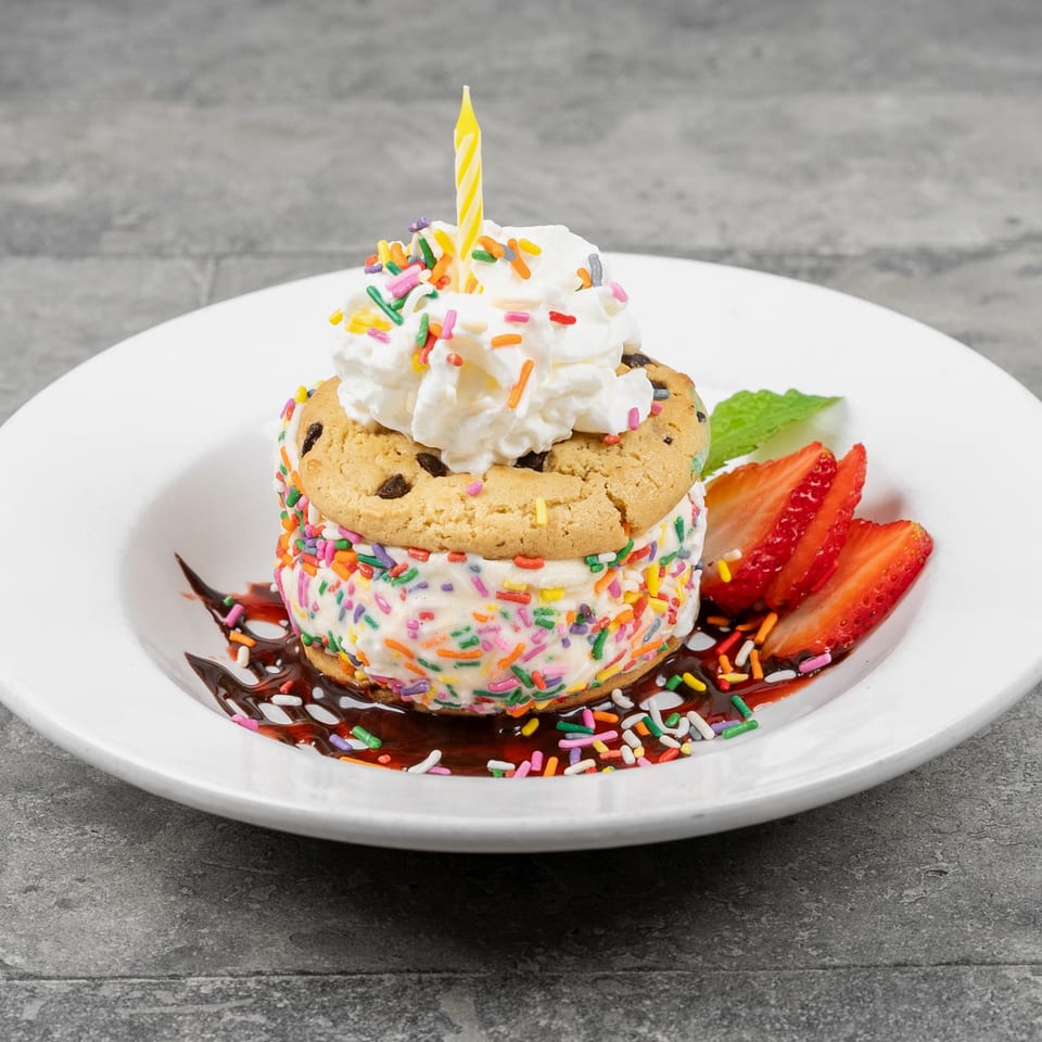 Rainbow-sprinkle cookie ice cream sandwich topped with whipped cream and candle, served with strawberries and chocolate drizzle.