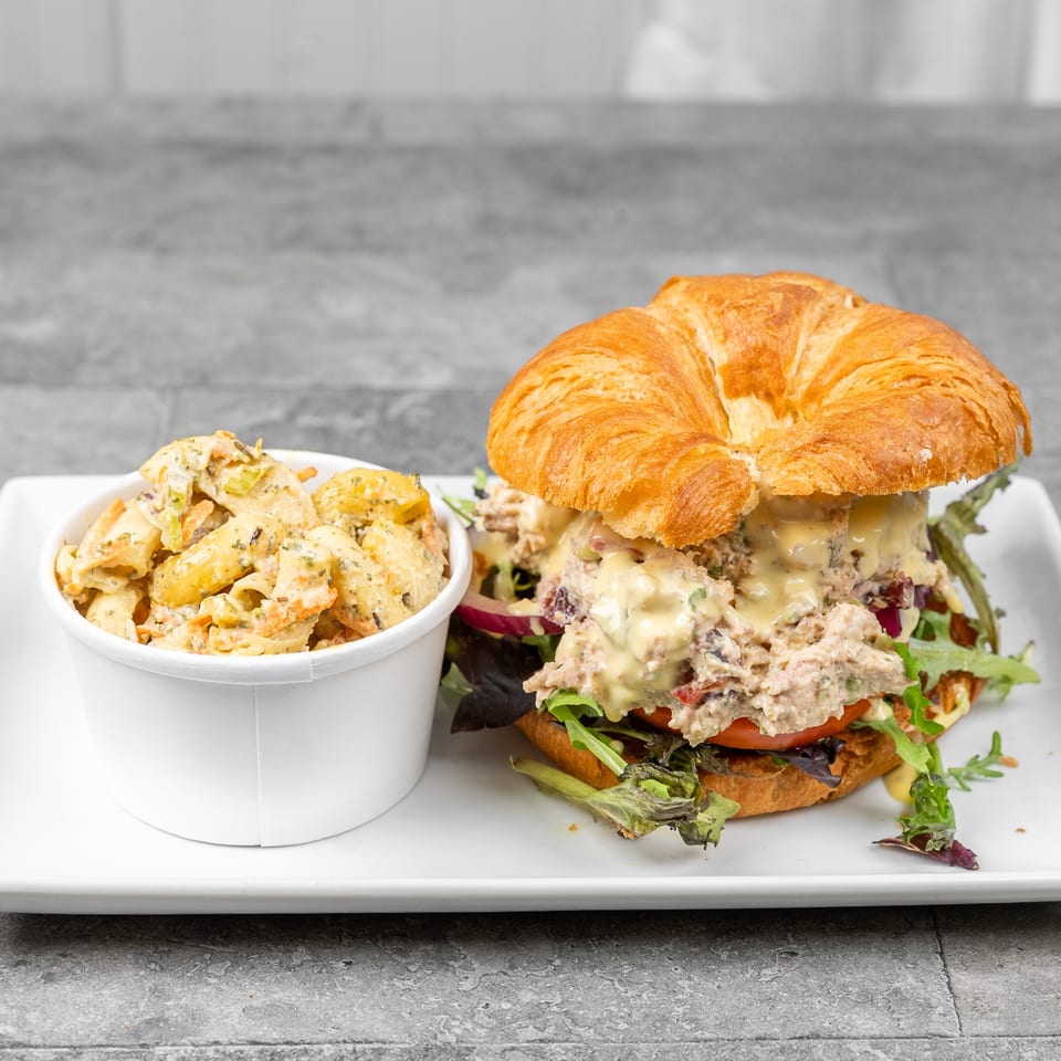 Croissant sandwich with creamy chicken salad, greens, tomato, and red onion served alongside pasta salad on a white plate.